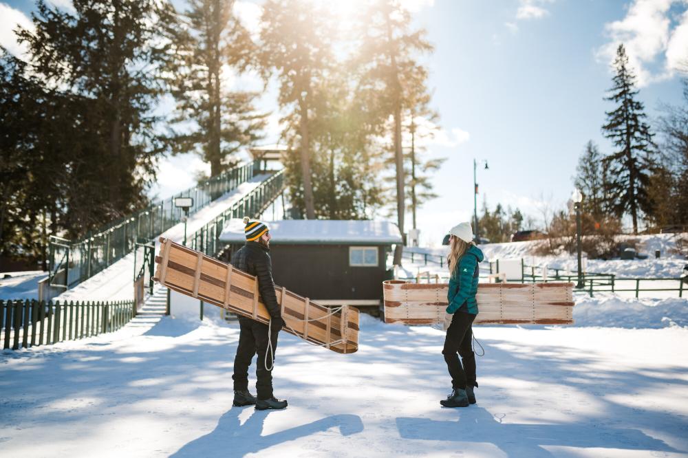 A man and woman chat on the ice while carrying toboggan sleds.