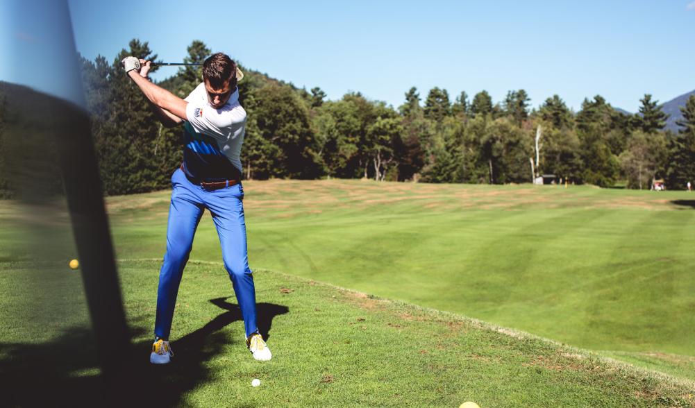 A man raises his club to hit a golf ball in front of a tree-line.