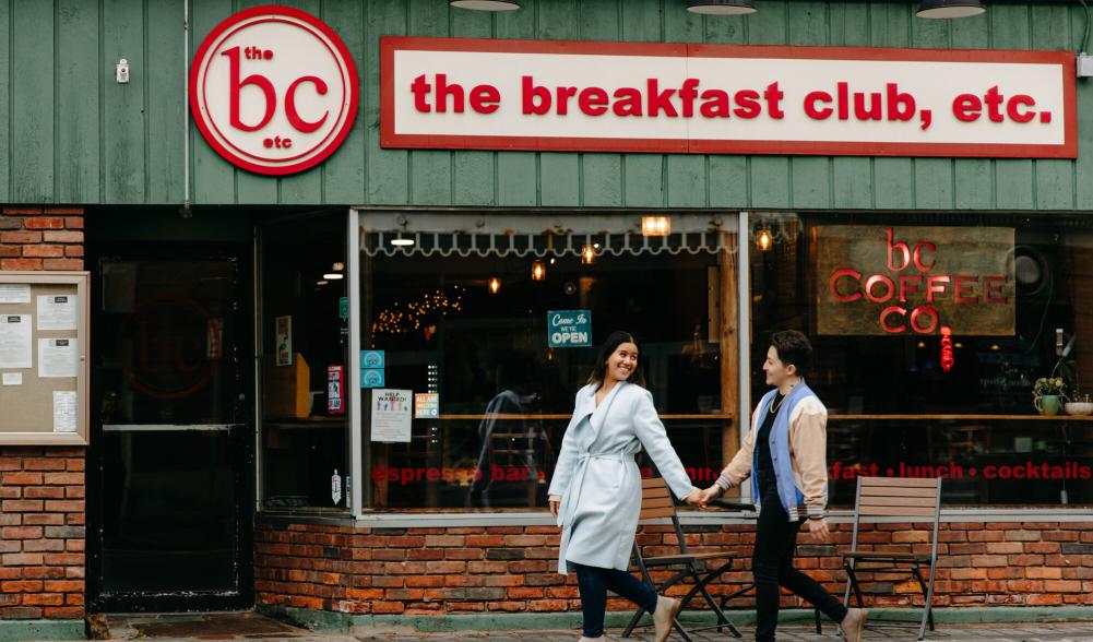 Two woman hold hands, one looking back over her shoulder at the other, walking toward the door of Breakfast Club restaurant