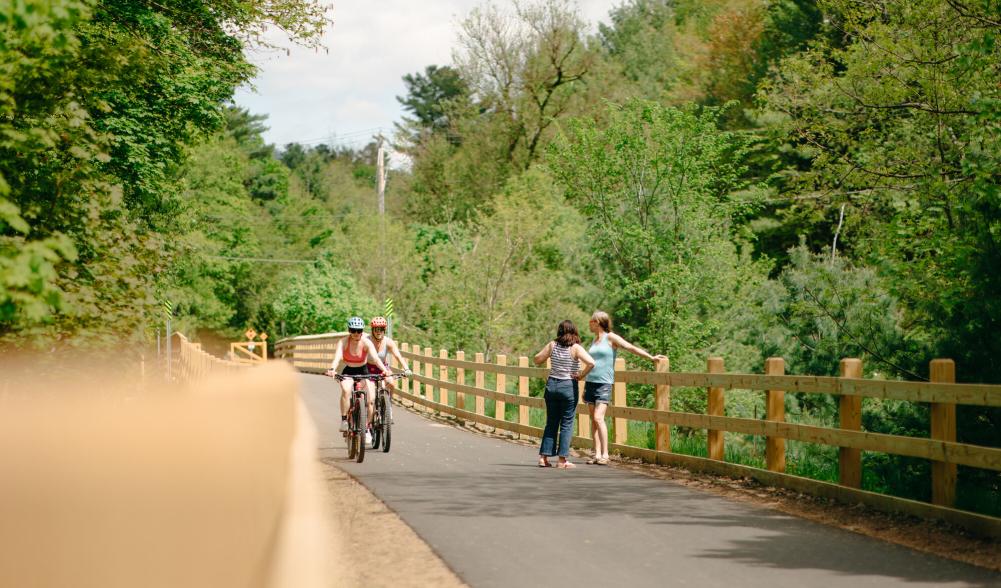 Two women stand on rail trail while bikers pass