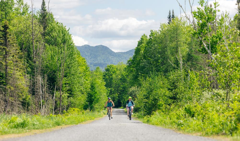 Two bikers ride side by side on rail trail with mountains in background