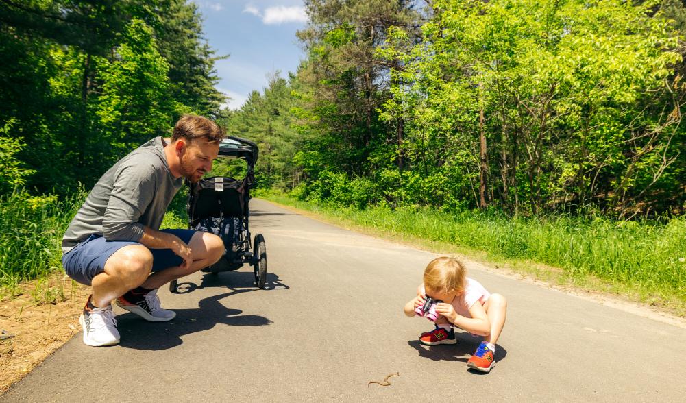 Father with stroller crouches next to small daughter crouched on rail trail looking at snake through binoculars