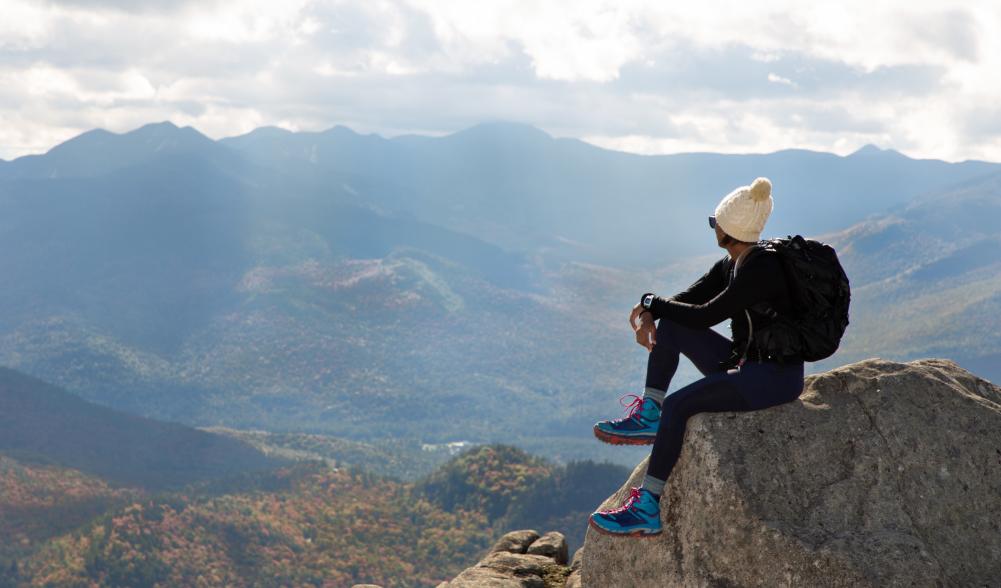 A woman sitting on a rock at the summit of a hike overlooking the mountains.