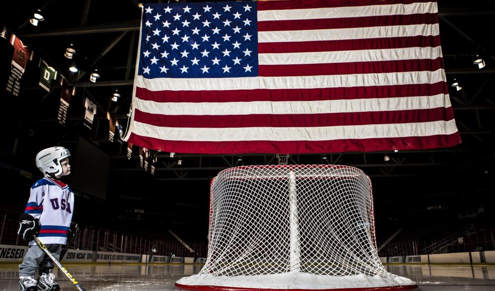 A small child in hockey gear looks upward at an American flag over a rink.