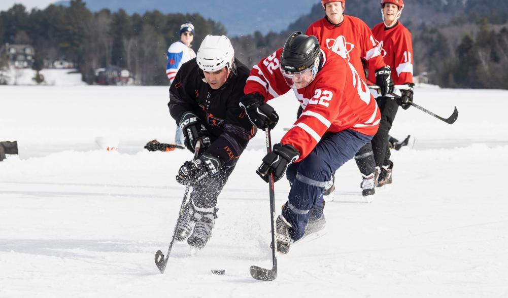 Two hockey players fight for control of the puck on a frozen lake on a sunny day.