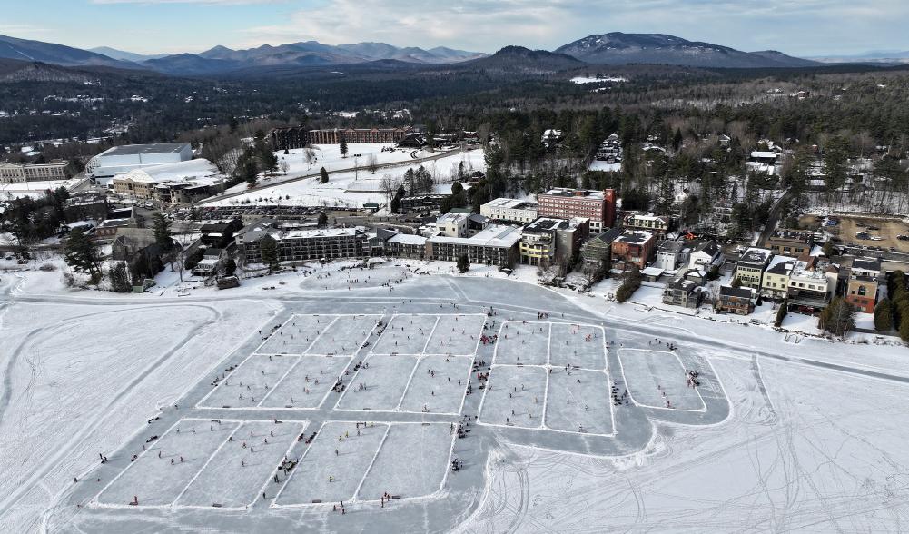 An aerial view of a frozen lake, divided into numerous pond hockey rinks. A small town overlooks the lake.