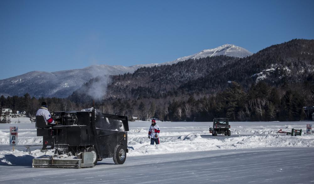 A zamboni driver sweeps across an outdoor rink with snowy mountains in the background.