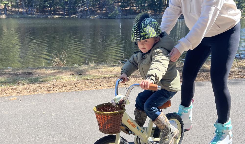 A woman on rollerblades guides a small child on a training bicycle.