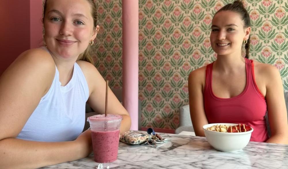 Two young women smile over a light lunch in a small cafe with pink and green decor.