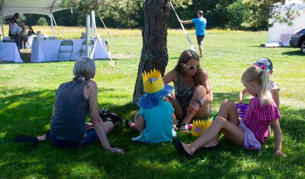 A woman sits under a tree eating snacks with her young kids.