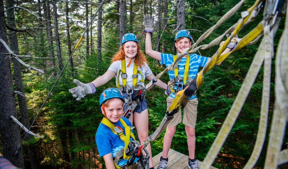 Three children smiling and geared up to go, posing for a picture at the zipline.