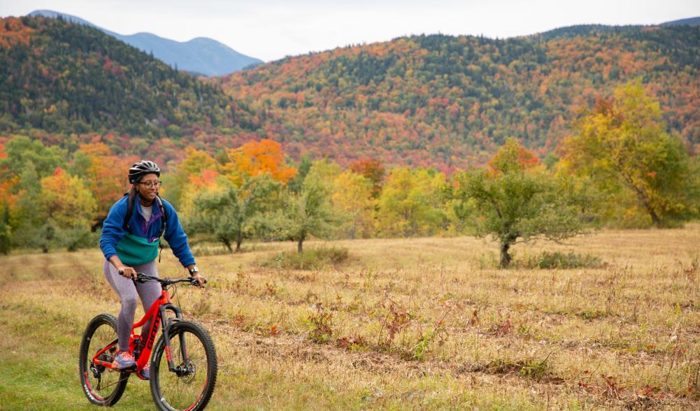 A woman mountain biking on a grassy trail with fall foliage in the background.