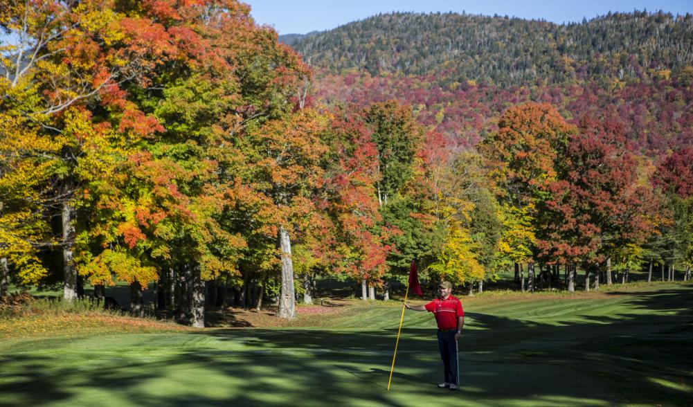 A man standing on the golf course holding a flag and smiling with beautiful fall leaves surrounding him.