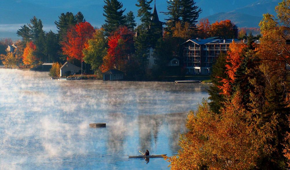 A person is paddling a canoe on Mirror Lake with morning mist coming off the water, fall trees surrounding them, and mountain views in the background.