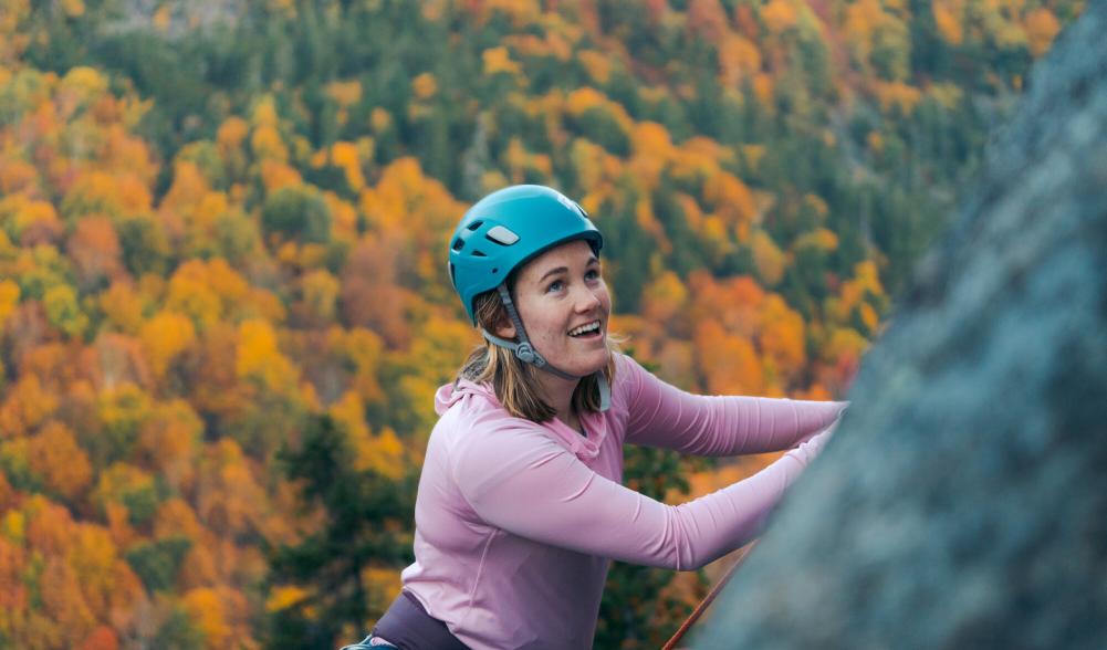 A woman smiling while rock climbing with an aerial view of the fall foliage below her.