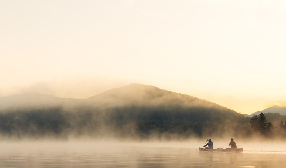 A canoe with two paddlers floats on mist-covered lake with mountain in background