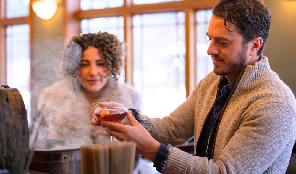 Man pulls cocktail from smoking box at bar as woman looks on