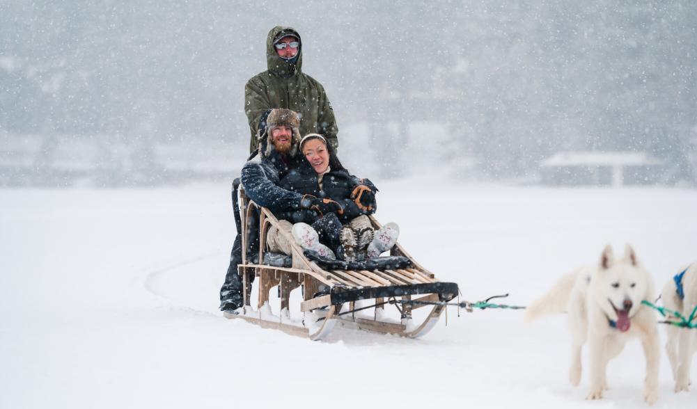 Man and woman sit in sled pulled over frozen lake by dog team