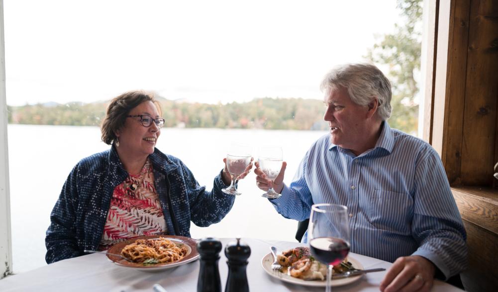 A couple clinking their beverages while dining at Jimmy's 21, seated in front a window with views of Mirror Lake.