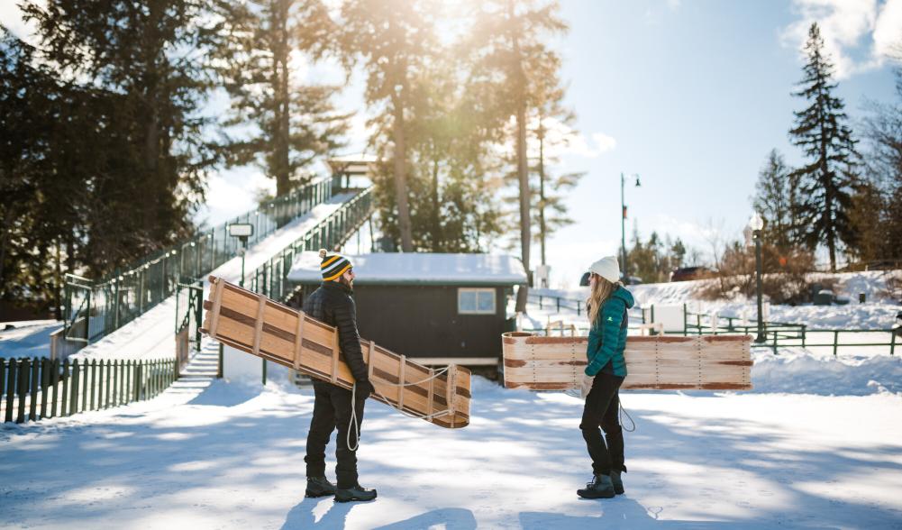 A man and woman chat on the ice while carrying toboggan sleds.