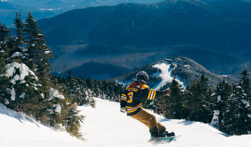 A woman in a bruins jersey snowboards down a mountain.