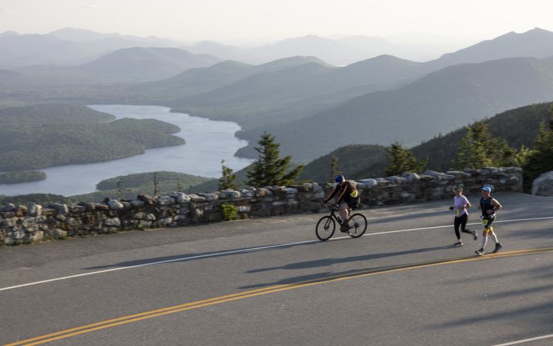 A cyclist and runners going up a road with mountain and lake views
