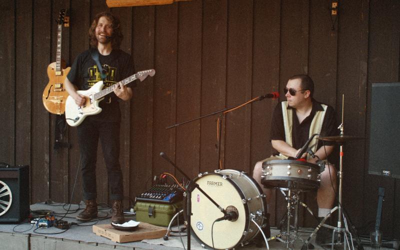 A music duo (man with guitar, a second man playing drums) plays on a patio.