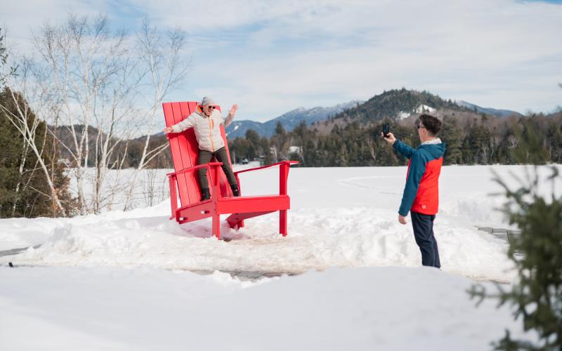 A man takes a photo of another man posing on a giant Adirondack chair in a snowy park with mountains in the background.