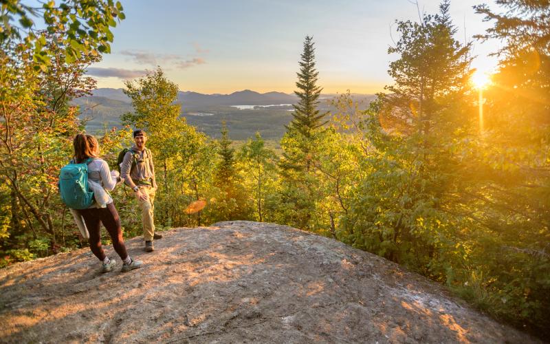 A couple talks to each other at the summit of Haystack Mountain while the sun is setting in the background