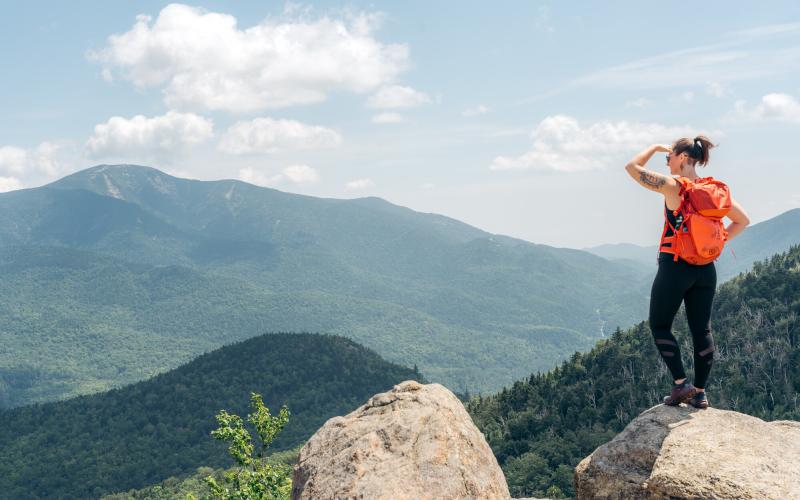 Hiker with red backpack shades eyes while looking at summit view on Rooster comb