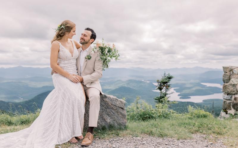 Bride sits on grooms lap atop a rock with Lake Placid in the background