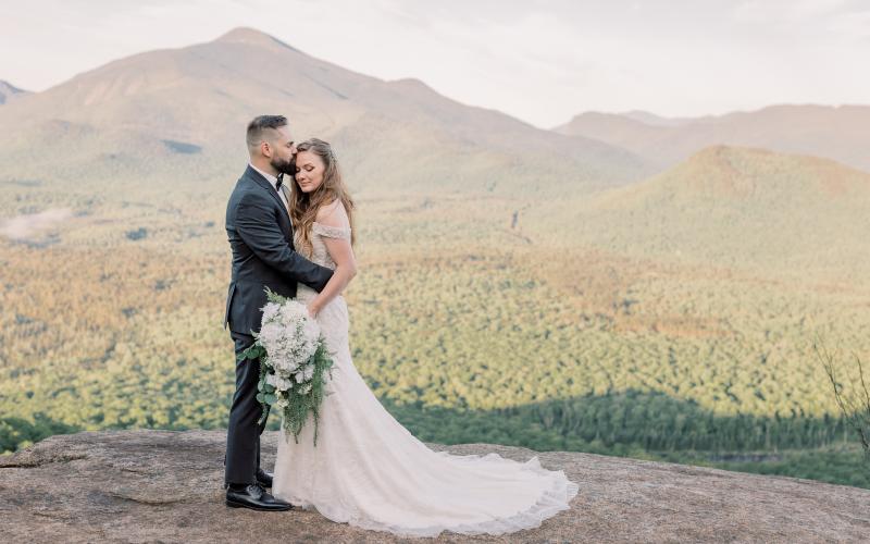 Groom kissses bride's forehead while she displays her bouquet on a rocky face with mountain views