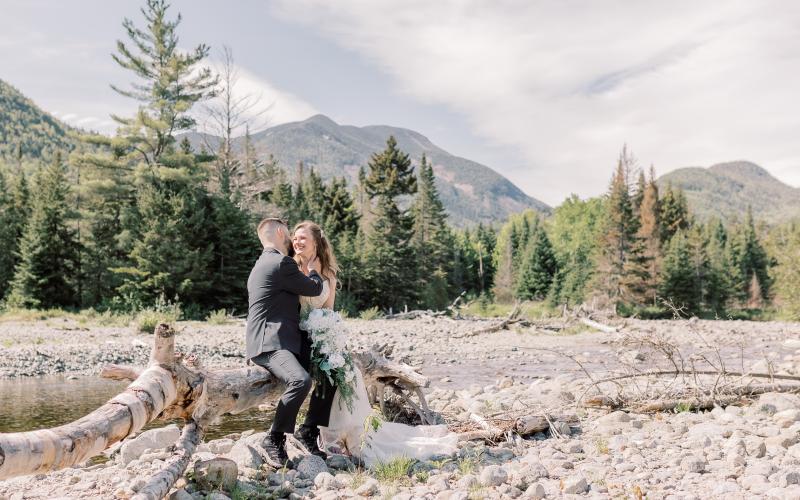 bride and groom snuggle on a fallen tree by a rocky river bed with mountain backdrop