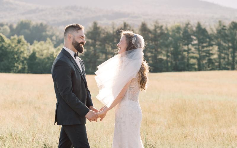 Bride and groom smile at each other in a wheaty field with veil blowing in the breeze