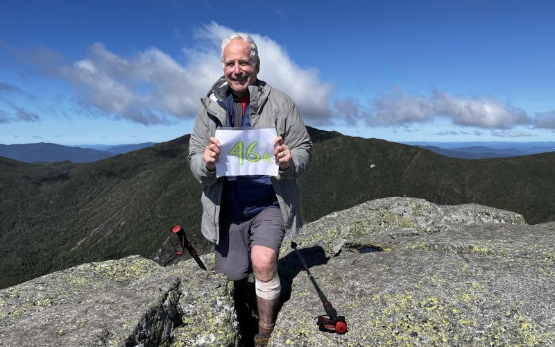 Older Gentleman stands atop a mountain peak holding a sign that reads '46er!'.