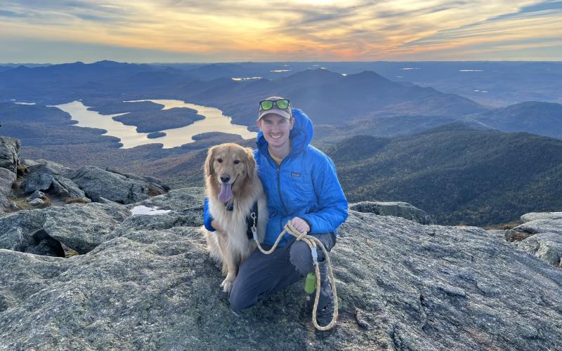 A man and his golden retriever sit atop a summit overlooking Lake Placid at sunset