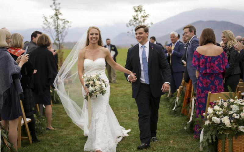 A bride and groom walk away from the alter while holding hands and smiling at friends and family and the mountain backdrop