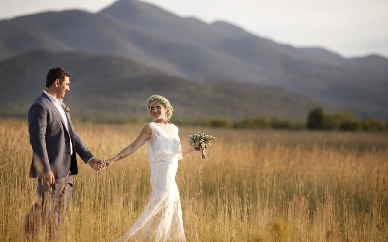 Bride and groom walk through a golden grass field with a mountain backdrop