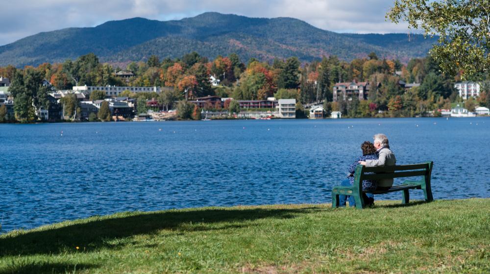 Older couple sits on a bench by Mirror Lake with Lake Placid and mountains in the background