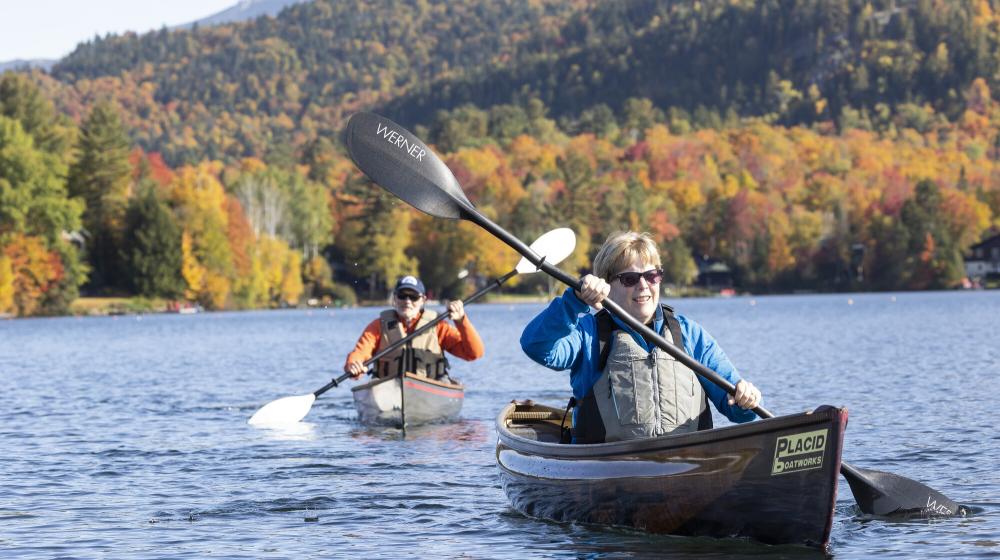 An older man and woman paddle on a lake during fall foliage.